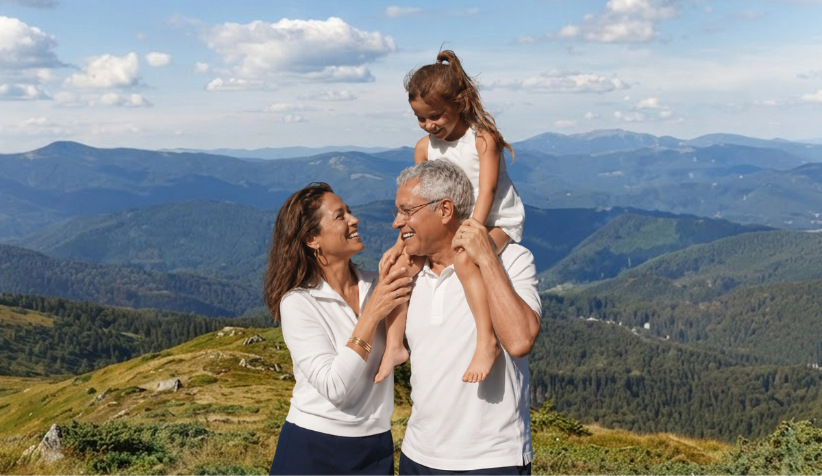 A smiling grandfather carries his granddaughter on his shoulders beside his wife on a mountain trail
